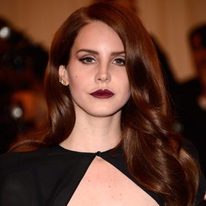 A person with long brown hair and dark lipstick stands, wearing a black outfit, against a blurred indoor background.