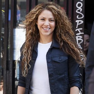 A woman smiles while wearing a dark jacket and white shirt, standing in front of a building with vertical bars and partially visible graffiti.