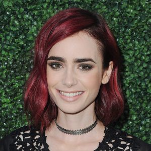 A person smiles against a lush green leafy background, wearing a black top and choker, with short, vibrant red hair.