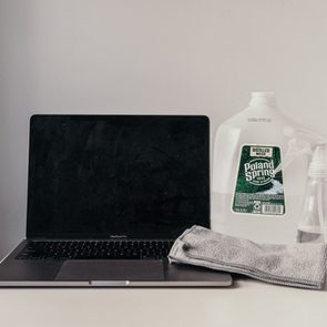 Laptop closed on table with cleaning cloth, spray bottle, and distilled water jug nearby in a simple, minimalistic setting.