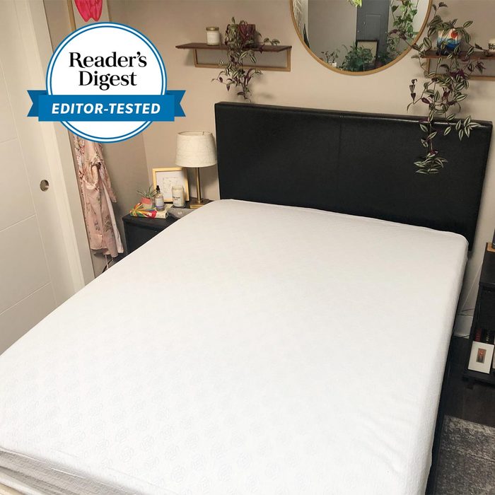 Bed with white sheet sits in a cozy room, surrounded by nightstands, plants, a mirror, and a lamp. 