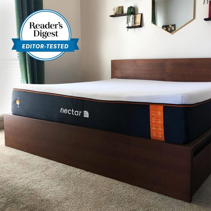 Mattress sits atop wooden bed frame, positioned in a carpeted room with wall decor and green curtains. Text: 