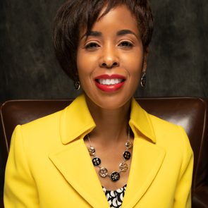 A person smiles, wearing a bright yellow jacket and necklace, seated in front of a dark background.