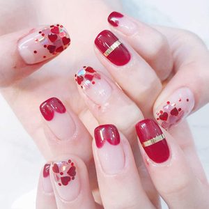 Fingernails showcase red nail art with decorative hearts and a metallic stripe, set against a light background.