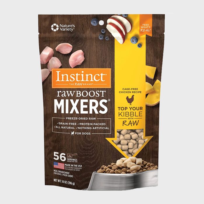 Dog food bag displays freeze-dried raw chicken mixers; scattered ingredients, text promotes grain-free, cage-free recipe; placed in kitchen-like setting.