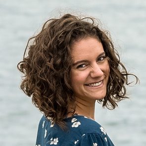 A woman with curly hair smiles, wearing a floral dress, against a blurred outdoor background.