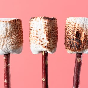 Three toasted marshmallows rest on sticks, against a pink background.
