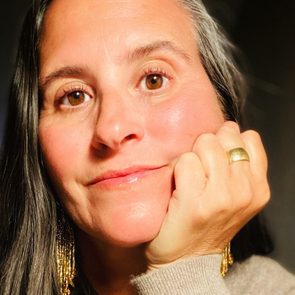 A person rests their chin on their hand, wearing a large ring and gold earrings, with a neutral expression, indoors.