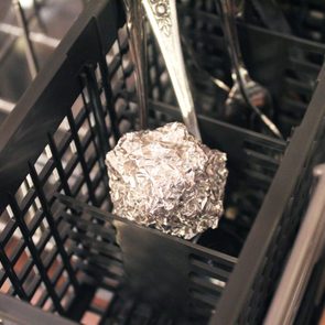 Aluminum foil ball sits in a dishwasher utensil rack among silverware, surrounded by dark plastic grid compartments.