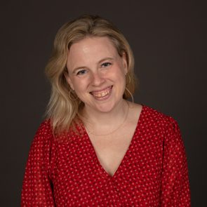 A smiling person wearing a red patterned blouse stands against a dark background.