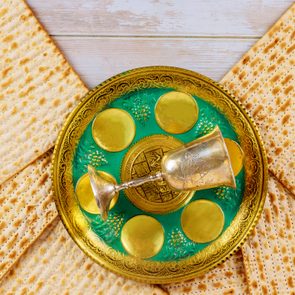 A silver goblet rests on an ornate, gold-edged Seder plate amid arranged matzah on a wooden table.