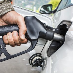 A hand grips a fuel nozzle, dispensing gasoline into a white car's tank at a gas station.