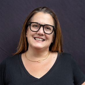 A person smiles, wearing glasses and a necklace, against a plain dark background.