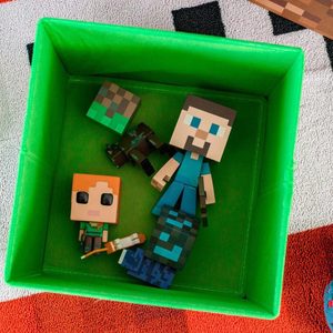 Toy figures lie inside a green box on a patterned fabric surface, depicting blocky, pixelated characters.