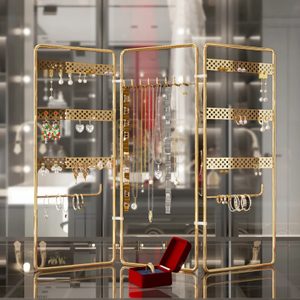 Jewelry stand displays earrings, necklaces, and bracelets; gold frame and perforated panels; set on a reflective surface with a red ring box in front.