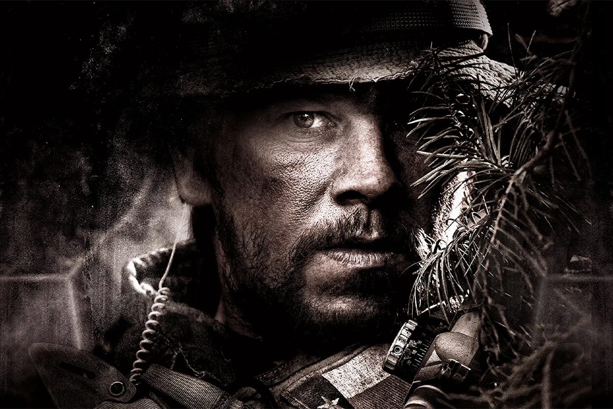 A rugged man wearing military gear peers through branches, focused and intense, in a dramatic and shadowy environment.