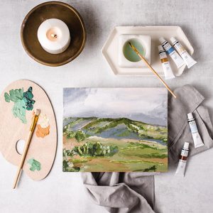 A landscape painting lies surrounded by paint tubes, brush, palette, candle, and cloth on a light surface, creating a serene artistic setup.