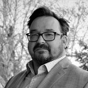 A person wearing glasses and a blazer stands outdoors, with leafless trees blurred in the background.