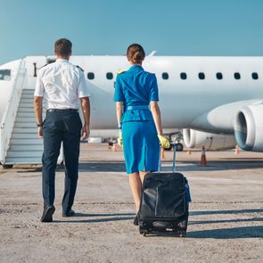 Pilot and flight attendant walk towards an airplane, carrying luggage, on an airport tarmac under a clear sky.