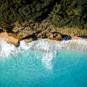 Waves crash against rocky shoreline, bordered by dense green foliage, forming a vivid marine landscape from an aerial viewpoint.