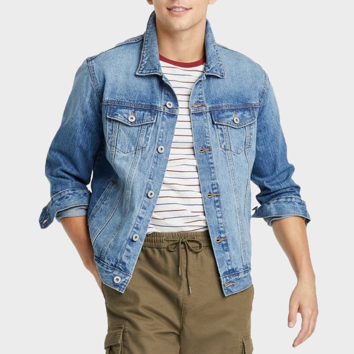 Man wears a denim jacket over a striped shirt, with hands in green cargo pants pockets, against a plain background.