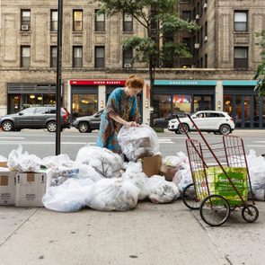 Individual searches through garbage bags on a city sidewalk, surrounded by buildings, parked cars, and a red shopping cart.