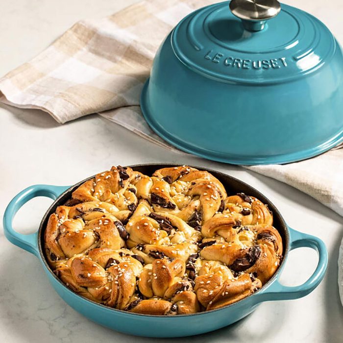 A baked pastry rests in a blue Le Creuset pan beside a matching lid on a checkered cloth on a marble surface.