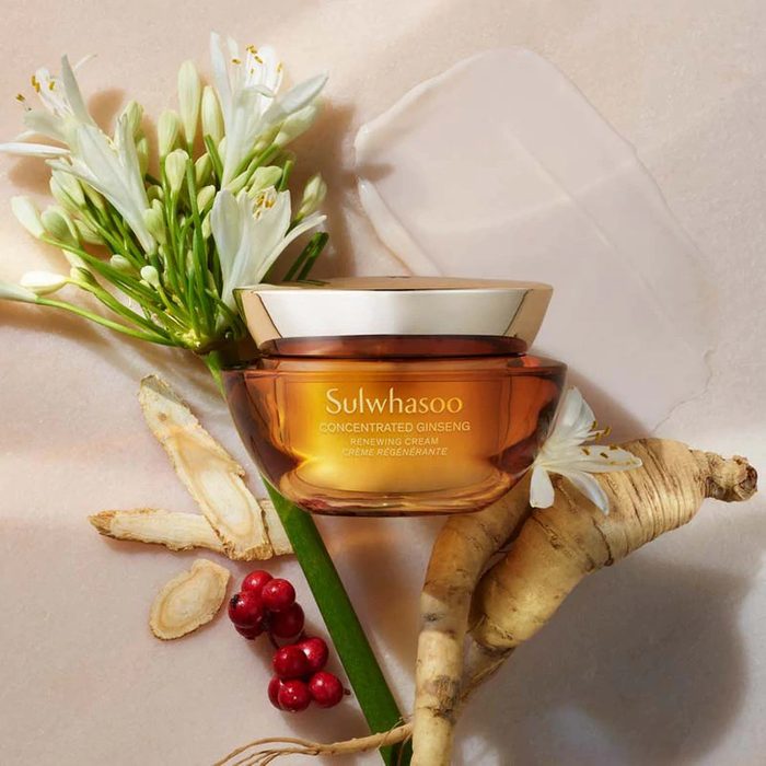 Cream jar sits with ginseng, flowers, and berries on beige surface. Text: 