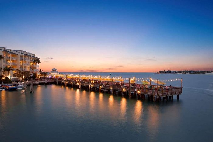 Pier with string lights glows softly at dusk, beside a calm ocean. Nearby, a building adds to the serene waterfront scene.