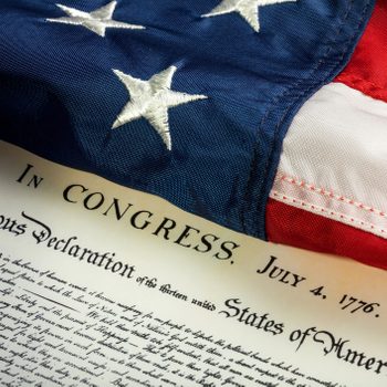 The American flag drapes over the Declaration of Independence document, dated July 4, 1776, in a symbolic historical setting.