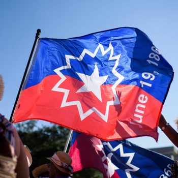 Flag flutters in the wind, displaying a star and date "June 19, 1865," amidst a sunny gathering of people outdoors.