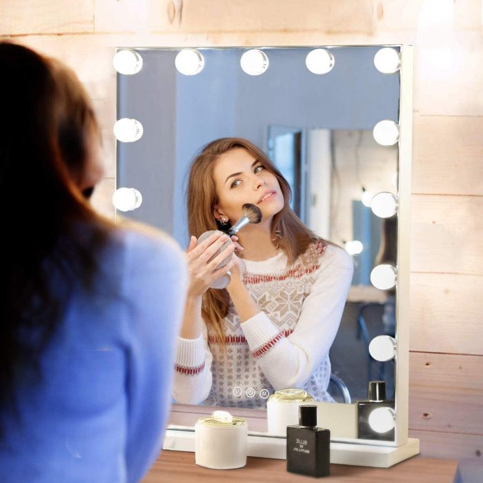 Woman applying makeup in front of illuminated vanity mirror, surrounded by wooden walls, with perfume bottles on the counter.
