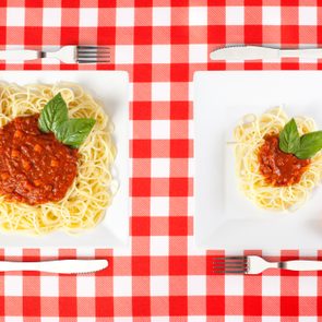 Two square plates hold spaghetti with tomato sauce and basil. Plates are on a red-and-white checkered tablecloth, accompanied by cutlery and sliced tomatoes.