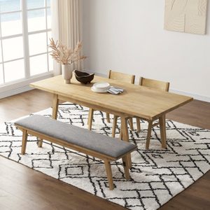 Wooden dining table displays plates and vase; set with chairs and bench; situated on patterned rug beside large window with ocean view.