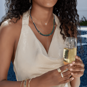 A woman in a light dress holds a champagne glass outdoors, adorned with layered necklaces and bracelets, near a tiled pool.