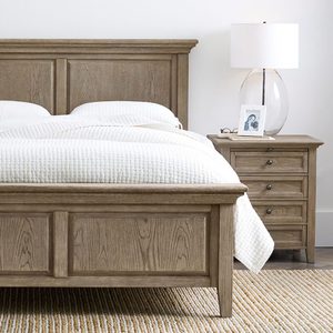 Wooden bed sits with white bedding; bedside table holds a lamp, framed photo, and eyeglasses on a striped rug in a bright room.