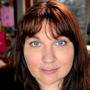A person with brown hair and blue eyes is smiling slightly in a room with colorful clothing visible in the background.