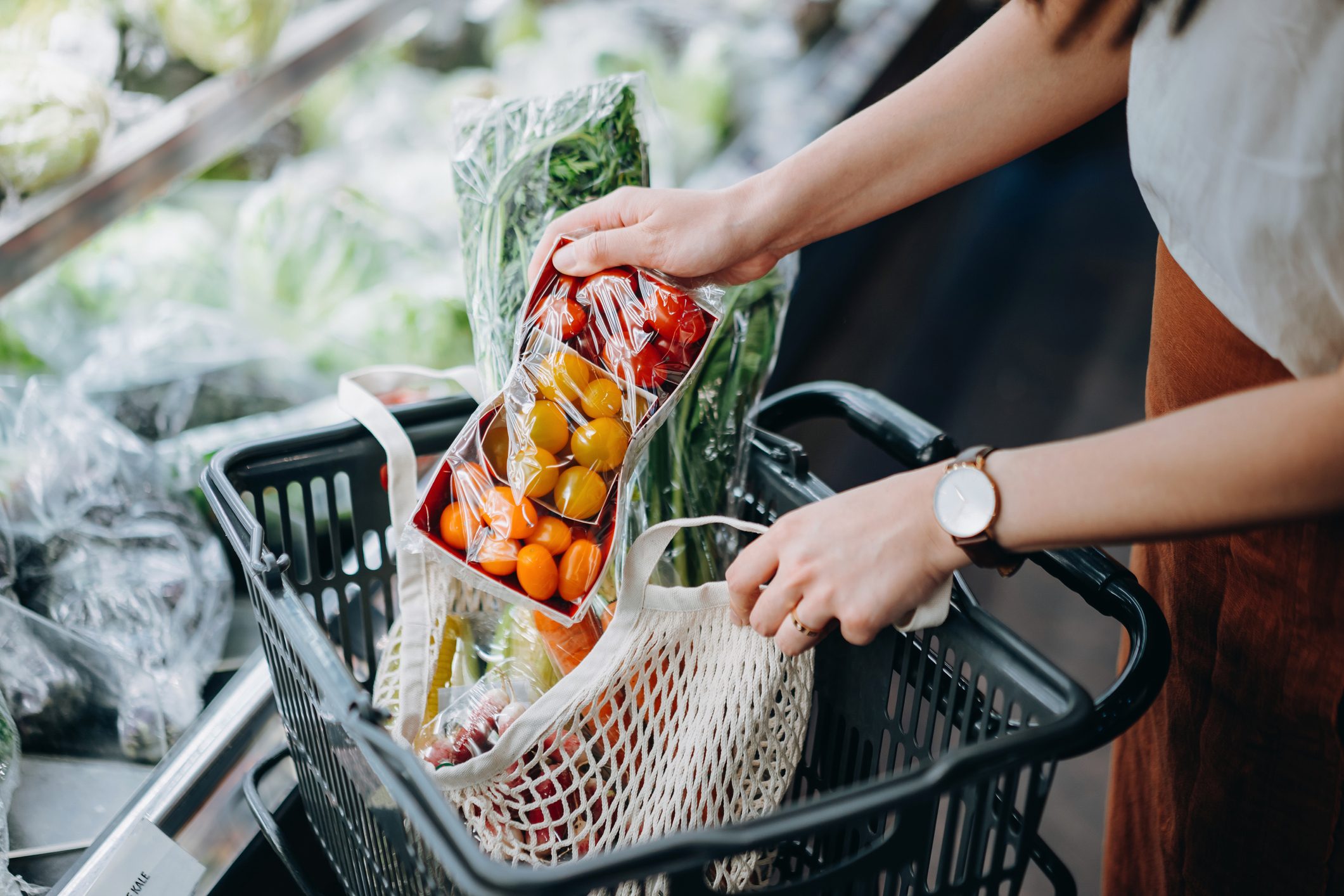 Cropped shot of young Asian woman shopping for fresh organic groceries in supermarket and putting a variety of fruits and vegetables into cotton mesh eco bag in a shopping cart. Zero waste concept