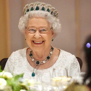 A smiling elderly woman with glasses wears an emerald tiara and necklace, seated at a table adorned with white flowers and glassware.