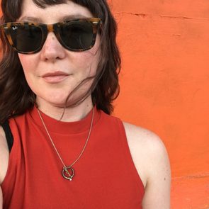 Person wearing sunglasses and red top against bright orange wall, with a necklace visible.
