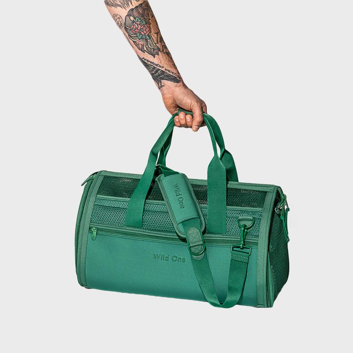 A green duffel bag is held by a tattooed arm, featuring a mesh panel and the text 