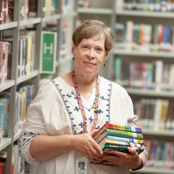 Person holds stack of books in a library aisle, surrounded by shelves filled with colorful books and green lettered signs.