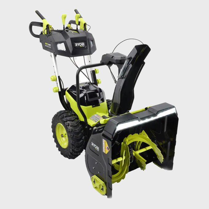 Snow blower resting on a flat surface, surrounded by a gray background. It features large wheels, green accents, and 