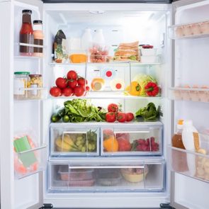 Refrigerator displaying various foods, including fruits, vegetables, juices, and condiments, arranged on shelves and inside drawers, in a well-lit kitchen setting.