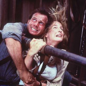 Two people cling tightly to a metal beam, wind blowing their hair, in an intense, turbulent environment.