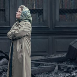 Person stands with arms crossed, wearing a coat and headscarf, amid scattered debris and rubble against a dark, paneled background.