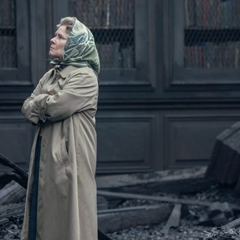 Person stands with arms crossed, wearing a coat and headscarf, amid scattered debris and rubble against a dark, paneled background.
