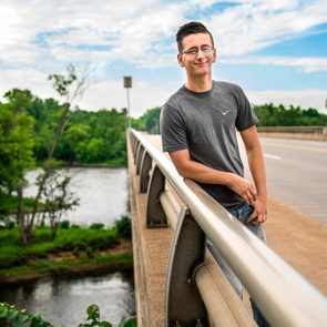 Person leans on bridge railing, wearing a gray shirt with a logo, surrounded by greenery and a river below, under a partly cloudy sky.