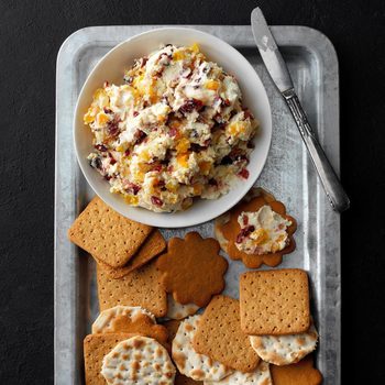 A bowl of fruit and cream cheese spread rests on a metal tray with various crackers around it, beside a butter knife.