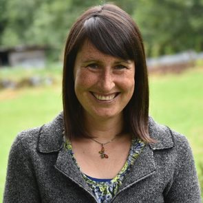 A person smiles outdoors, wearing a gray coat and patterned blouse, with a grassy field and trees in the background.